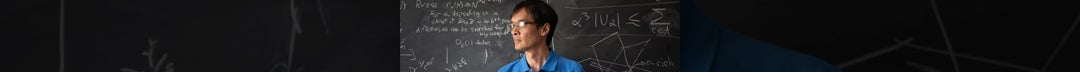 Terence Tao wearing a blue shirt and glasses stands against a solid chalkboard