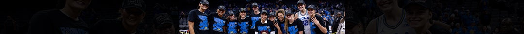 A group of young women stand in the middle of a basketball stadium, smiling, celebrating, and surrounding a trophy. The stadium is filled with people, confetti scattered on the floor, and the women are wearing shirts that read "We are not done".