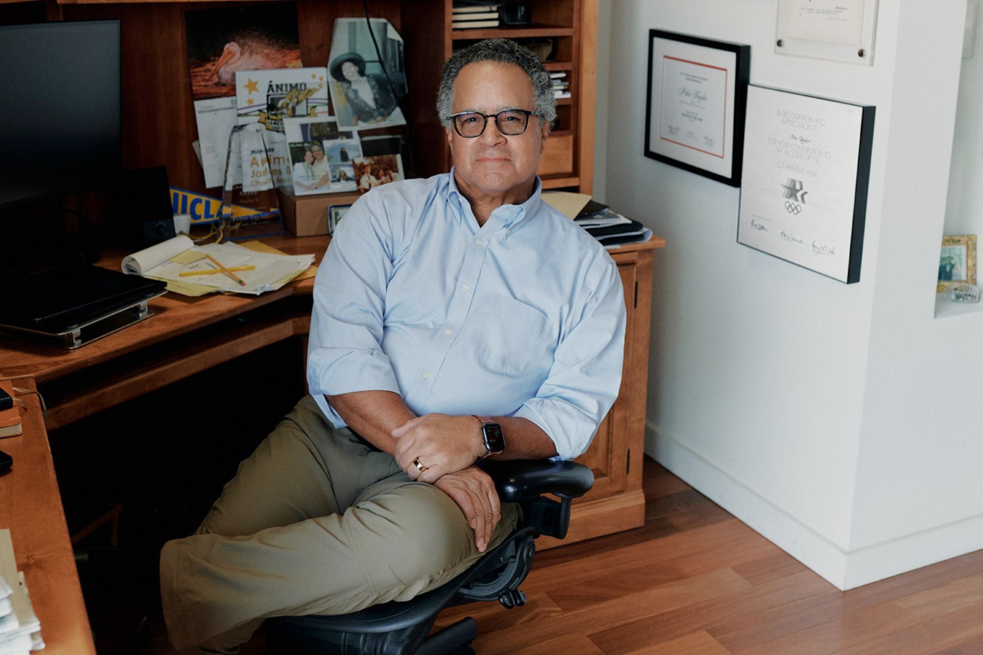 A man sits in an office chair with his legs crossed in a home office, surrounded by wooden cabinets, framed certificates and photos on the wall, and papers and notebooks on the desk.
