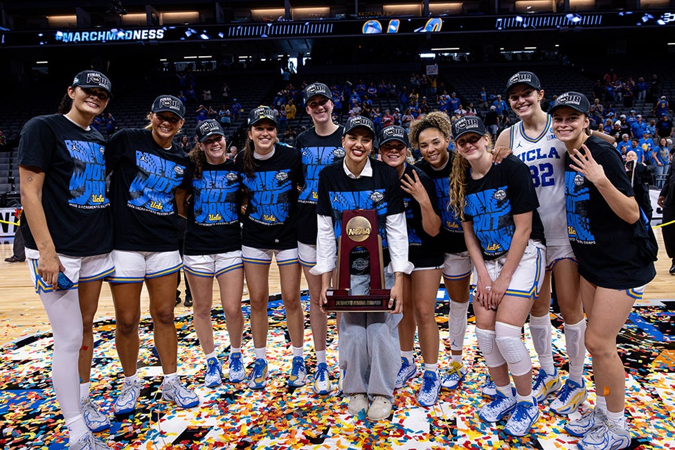 A group of young women stand in the middle of a basketball stadium, smiling, celebrating, and surrounding a trophy. The stadium is filled with people, confetti scattered on the floor, and the women are wearing shirts that read "We are not done, UCLA".