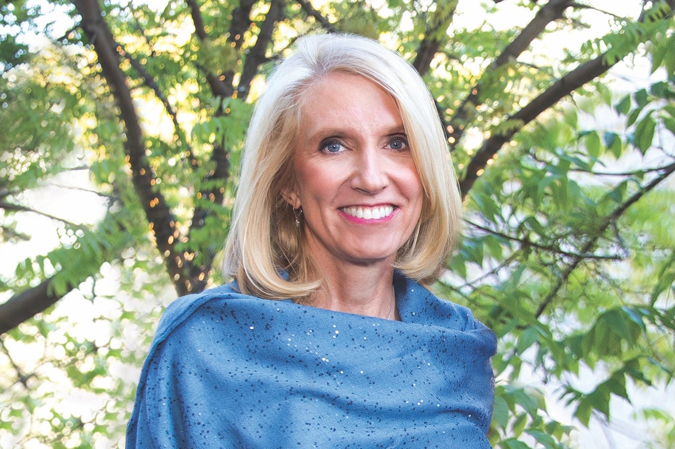 A woman with short blonde hair smiles in a blue wrap with trees in the background.