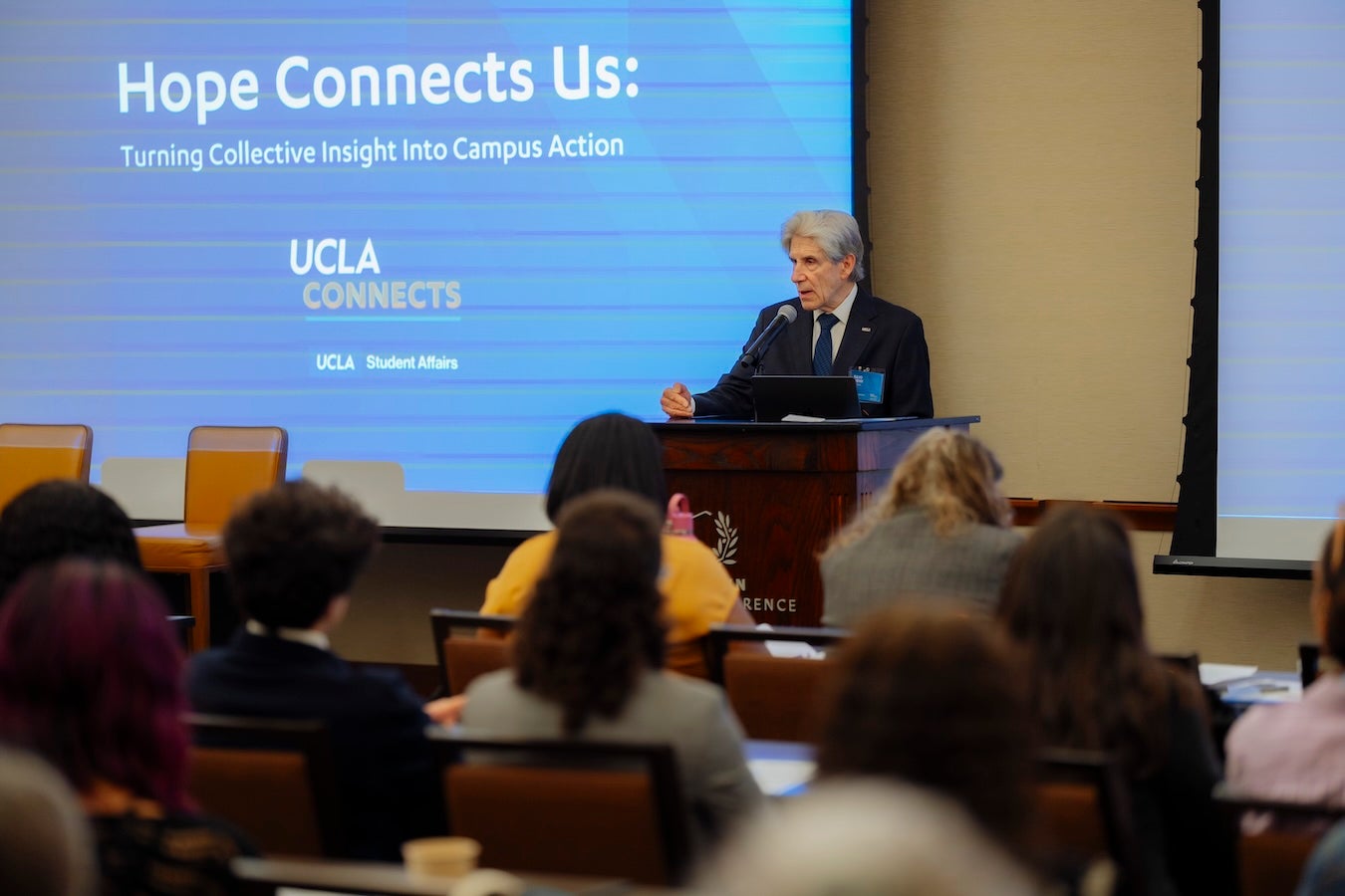 A man stands at a podium with an audience seated in front of him. He wears a blue suit and a projector is behind him with a slide for "Hope Connect Us". 